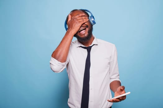 Cheerful man holding smartphone, palm over eyes, randomly selecting song from playlist to listen in wireless headphones. Facepalming freelancer listening to funny podcast through his headset.