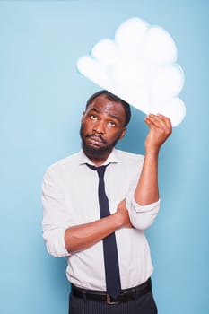 Portrait of male entrepreneur in white shirt looking up at white paper idea cloud brainstorming while standing in front of blue background. African American man thinking under thought bubble.