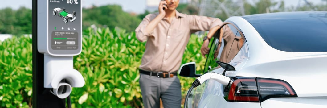 Man talking on the phone while recharge EV car battery at charging station connected to power grid tower electrical as electrical industry for eco friendly car utilization. Panorama Expedient