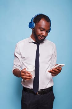 Young black man with wireless headphones using smartphone to browse social media while holding a warm drink. On his break, office worker listens to online music while drinking a cup of coffee.