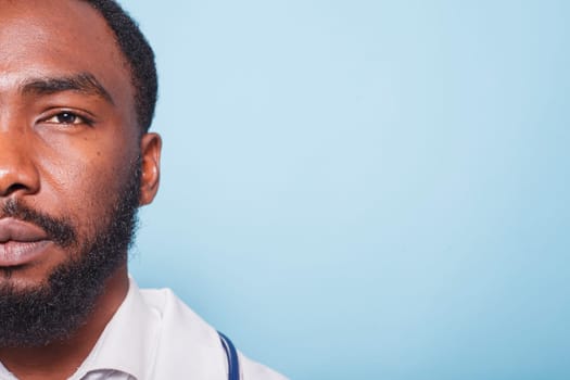Close-up of African American doctor with a beard looking at the camera. Half-face portrait of black male healthcare specialist wearing white lab coat against isolated blue background.