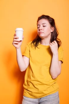 Tired woman posing against isolated orange background in casual clothes, holding coffee cup, relaxing with closed eyes. Sleepy expression of young brunette lady depicts exhaustion and weariness.