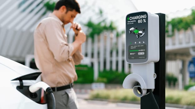 Young man put EV charger to recharge electric car's battery from charging station in city commercial parking lot. Rechargeable EV car for sustainable environmental friendly urban travel. Expedient