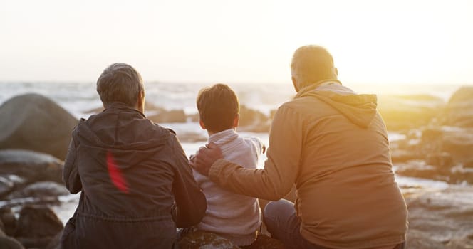 Grandparents, child and beach for sunset relax on rocks with back for connection, bonding or travel. People, kid and sitting together with rear view for ocean horizon for family, vacation or love.