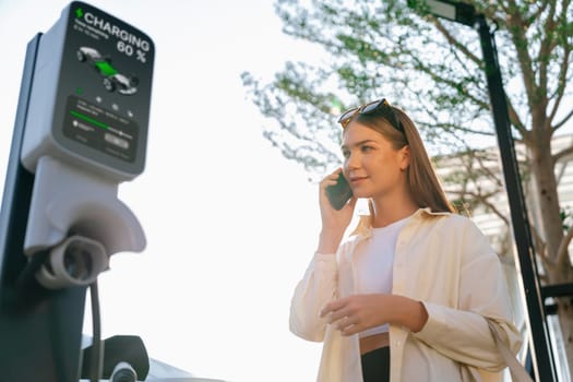 Young woman holding shopping bag talking on the phone while recharging EV car battery from charging station at city mall parking lot. Modern woman go shopping by eco car. Expedient