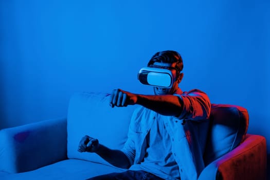 Happy man drive a car movement while wear VR glasses and sitting at sofa with neon light background. Smart gamer playing sport game while moving driving car gesture by using smart goggle. Deviation.