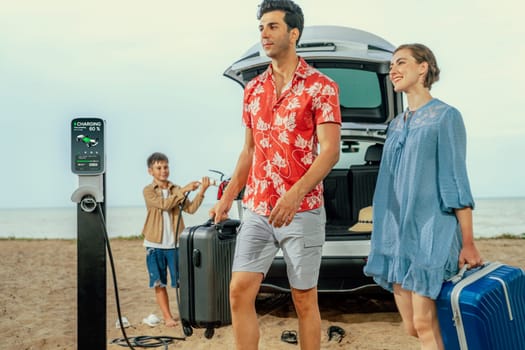 Family vacation trip traveling by the beach with electric car, lovely family taking luggage out while charging EV car battery with clean energy. Alternative family travel by eco-friendly car.Perpetual