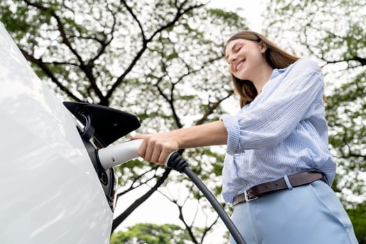 Young woman recharging battery for electric car during road trip travel EV car in natural forest or national park. Eco friendly travel during vacation and holiday. Exalt