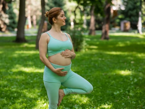 Pregnant caucasian woman doing yoga in the park. Tree pose