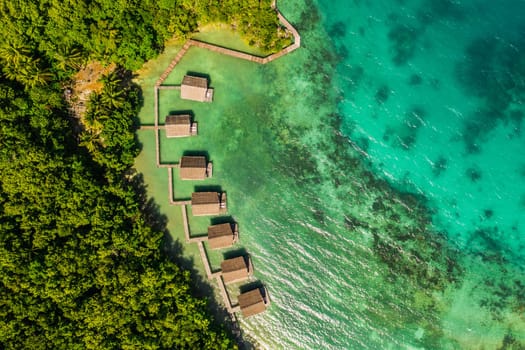 Aerial, buildings and lodge by tropical sea with bridge, island and trees on beach for tourism agency. Jetty, hotel and outdoor with ocean, hospitality and drone in nature with waves in Raja Ampat.
