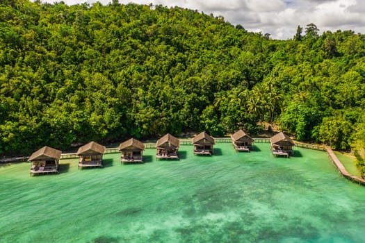 Aerial, bungalow and resort by tropical ocean with hill, island and trees on beach for tourism agency. Jetty, hotel and hospitality by sea, adventure and drone in nature with water in Raja Ampat.