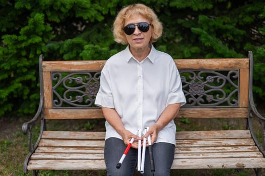 An elderly blind woman sits on a bench in the park with a folded tactile cane in her hands