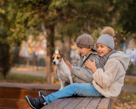 Brother and sister sit in an embrace with a dog on a bench for a walk in the autumn park. Boy, girl and jack russell terrier