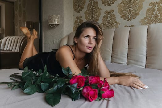 A woman lays on a bed with a bouquet of red roses in her lap. Concept of romance and intimacy, as the woman is surrounded by the flowers and he is in a vulnerable position