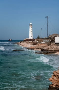 A lighthouse is on a rocky shoreline next to the ocean. The water is choppy and the sky is clear