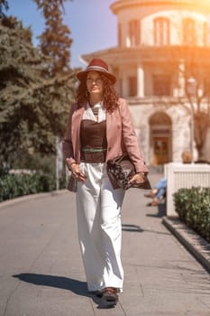 Woman park city. Stylish woman in a hat walks in a park in the city. Dressed in white corset trousers and a pink jacket with a bag in her hands