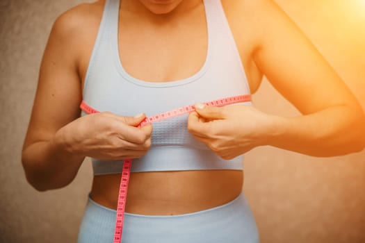 Cropped view of slim woman measuring breasts with tape measure at home, close up. Unrecognizable European woman checks the result of a weight loss diet or liposuction indoors. Healthy lifestyle..