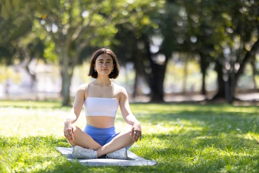 A woman is sitting on a mat in a park, looking up at the sky. She is in a peaceful and relaxed state