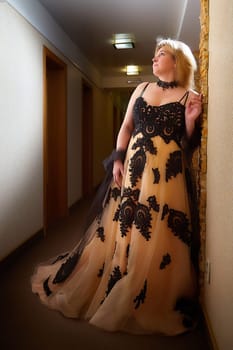 Full length view of a blonde female model, dressed in a black and white queen dress in dark corridor. Mysterious stranger alone inside castle or hotel