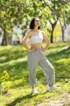 A woman is standing in a park, wearing a white tank top and grey sweatpants. She is looking up at the sky, and her posture suggests that she is feeling confident and relaxed