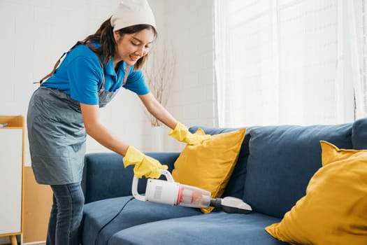 In a cozy setting an Asian woman focuses on housework using a vacuum machine cleaner to clean a sofa. Her dedication to hygiene and furniture care exemplifies a modern approach to cleaning.