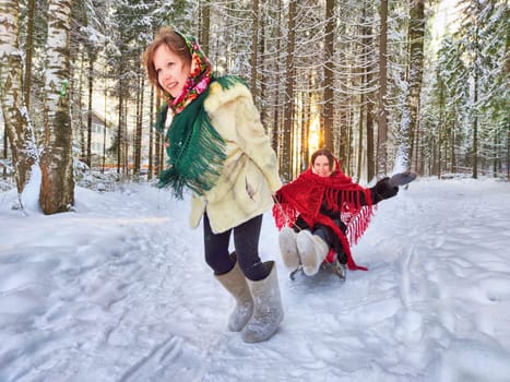 Mother and daughter in shawls are sledding in the winter forest. Ethnic clothes and fun on Shrovetide carnival Maslenitsa