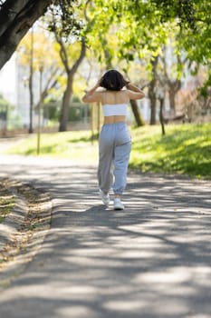 A woman is walking down a path in a park. She is wearing headphones and has her hands on her head. The scene is peaceful and serene, with the woman enjoying her walk in the park