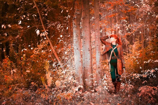 Mature model portraying a royal huntress with red curve hair is hunting with a crossbow in the in vibrant autumn forest in thematic photo shoot