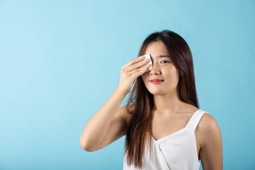 Facial Beauty Treatment Hygiene skin. Asian beautiful young woman removing cosmetic make up using pad cotton face cleaner disc, studio shot isolated on blue background, Happy female cleaning eyes