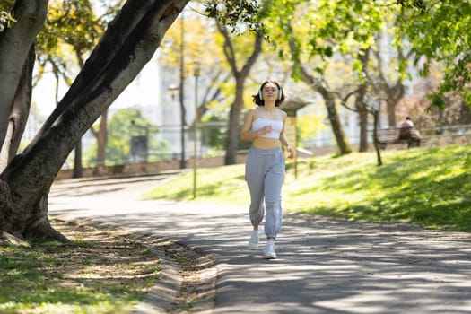 A woman runs on a path in a park. She is wearing headphones and a white tank top