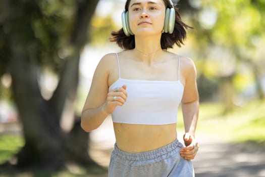 A woman is running in a park with her headphones on. She is wearing a white tank top and gray shorts