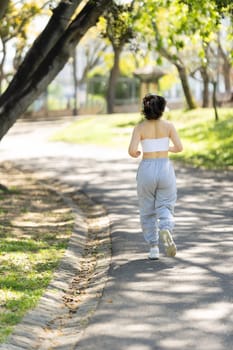 A woman is running on a path in a park. She is wearing a white tank top and gray sweatpants