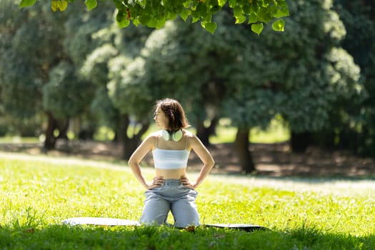 A woman is sitting on the grass in a park. She is wearing a white tank top and grey pants