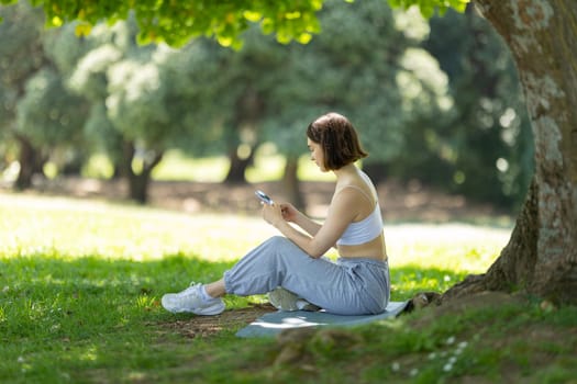 A woman is sitting under a tree and looking at her phone. She is wearing a white tank top and gray pants