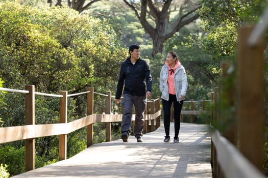 A man and woman are walking on a wooden bridge. The man is wearing a black jacket and the woman is wearing a pink jacket. They are holding hands and seem to be enjoying each other's company