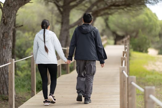 A man and woman are walking on a wooden bridge. The man is wearing a black jacket and the woman is wearing a gray jacket. They are holding hands and walking together