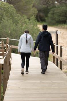 A man and woman are walking on a wooden bridge - rear view. The man is wearing a black jacket and the woman is wearing a pink jacket. They are holding hands and seem to be enjoying each other's company