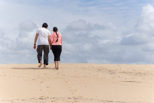 A couple is walking in desert, holding hands. The sky is cloudy, and the beach is sandy