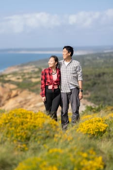A couple standing on a hillside with a beautiful view of the ocean. The man is wearing a plaid shirt and the woman is wearing a red plaid shirt