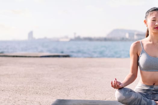 young asian woman in sportswear doing meditation by the sea sitting with legs crossed, concept of mental relaxation and healthy lifestyle, copy space for text