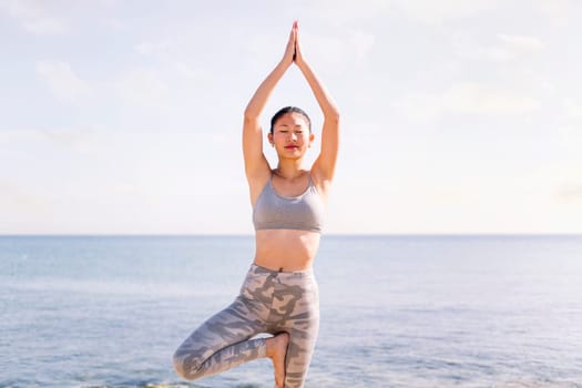 young asian woman in sportswear doing yoga tree position at beach, concept of mental relaxation and healthy lifestyle