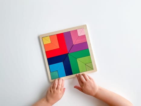 child's hand collects multicolored wooden mosaic on white background. child solves a colorful tangram. square of colorful geometric shapes on white background.