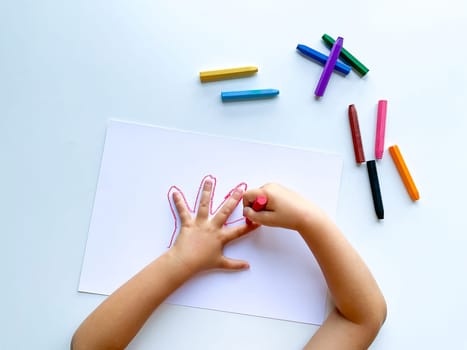 Childrens hands draw their hand with wax crayons on white paper, top view. High quality photo