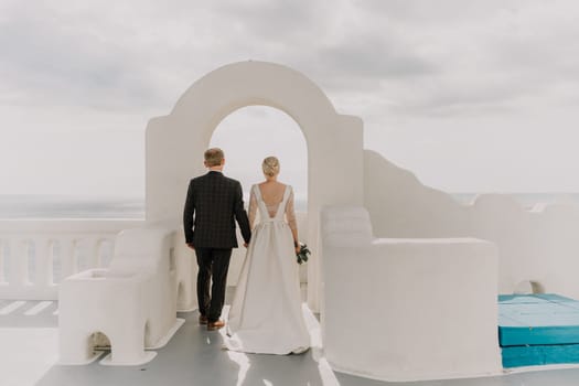 A bride and groom are walking through a white archway on a sunny day. The bride is wearing a white dress and the groom is wearing a suit. The archway is decorated with flowers