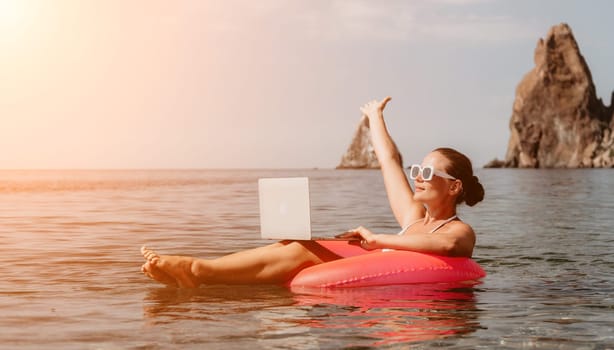 Woman freelancer works on laptop swimming in sea on pink inflatable ring. Pretty lady typing on computer while floating in the sea on inflatable donut at sunset. Freelance, remote work on vacation