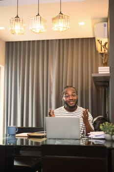 Cheerful African American man having video call on laptop sitting at table in kitchen.