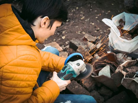 A young caucasian man in a yellow jacket is holding a drill and polishing with a disc in the backyard of a house with construction debris on the ground, close-up side view. The concept of the polishing process.