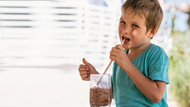 Happy childhood. Boy drinking chocolate tea with ice in hot summer day time. Cute lover of sweets and tasty things. Shows thumbs up.