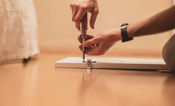 Young caucasian unrecognizable brunette man hand tightening a screw with a screwdriver on a white wooden wardrobe door while sitting on the floor, close up side view with selective focus. Furniture assembly concept.