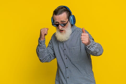 Excited senior stylish old man listening music via headphones and dancing disco party fooling around having fun expressive gesticulating hands. Happy elderly grandfather pensioner on yellow background